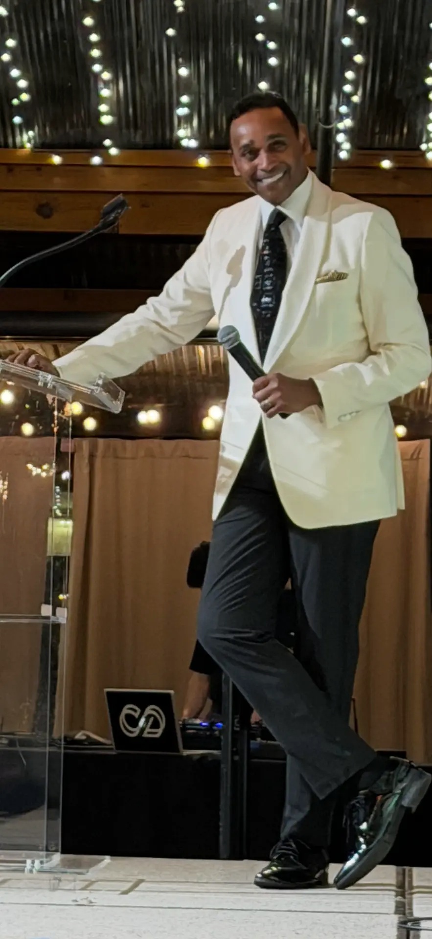Dwayne Ballen hosting an event in an ivory jacket, holding a microphone beside a transparent lectern