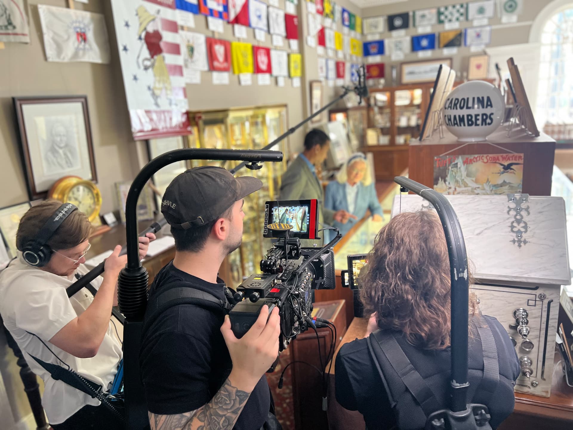 A film crew operating a cinema camera and boom microphone filming an interview in a golf shop, with a director's monitor in frame