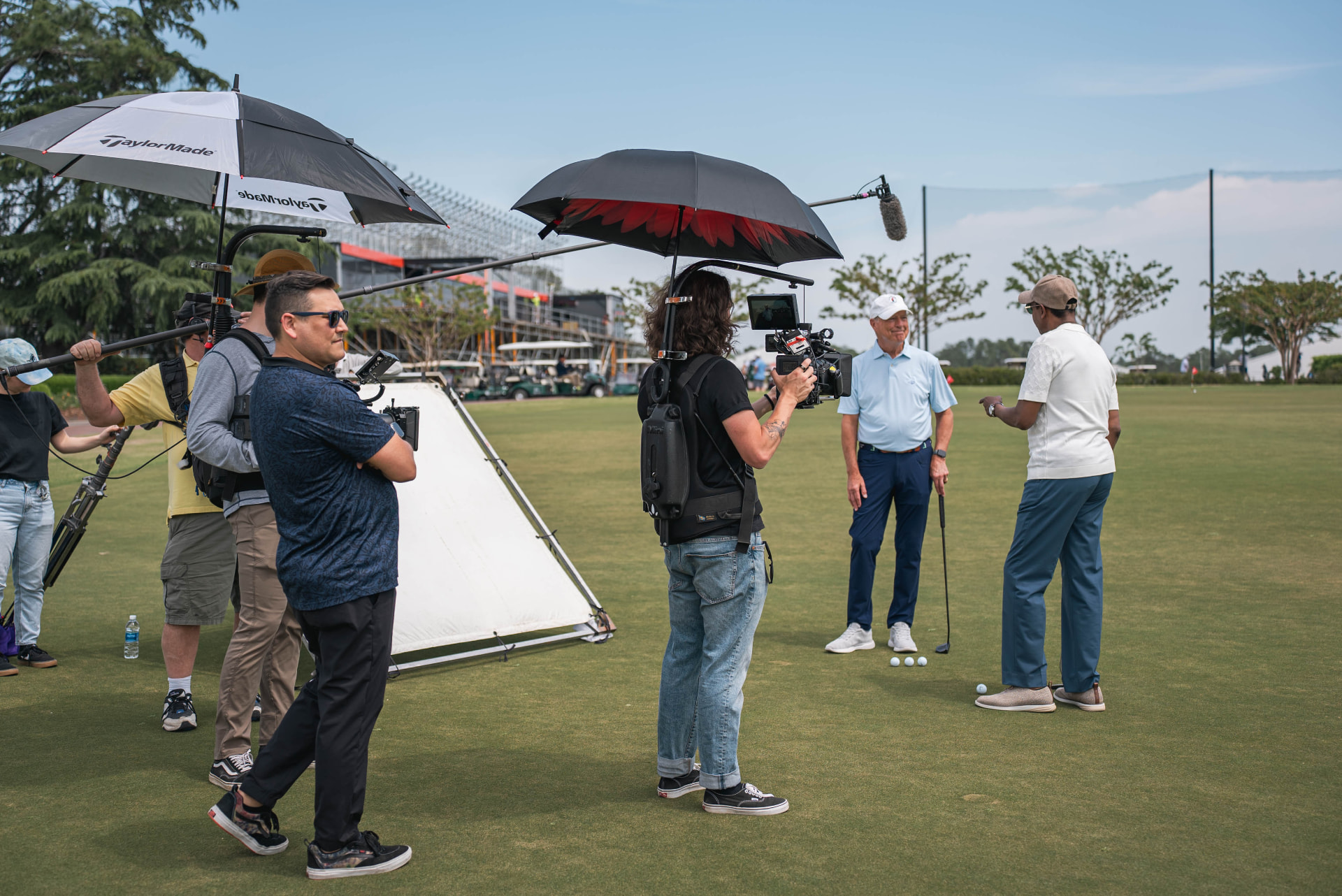A documentary crew filming an on-camera interview on a golf course under umbrellas, with a boom operator raising the microphone