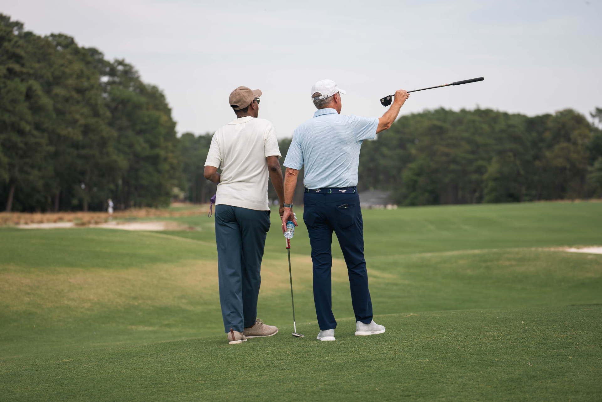Two figures walking across a golf course green during a PBS documentary production, one holding a golf club aloft