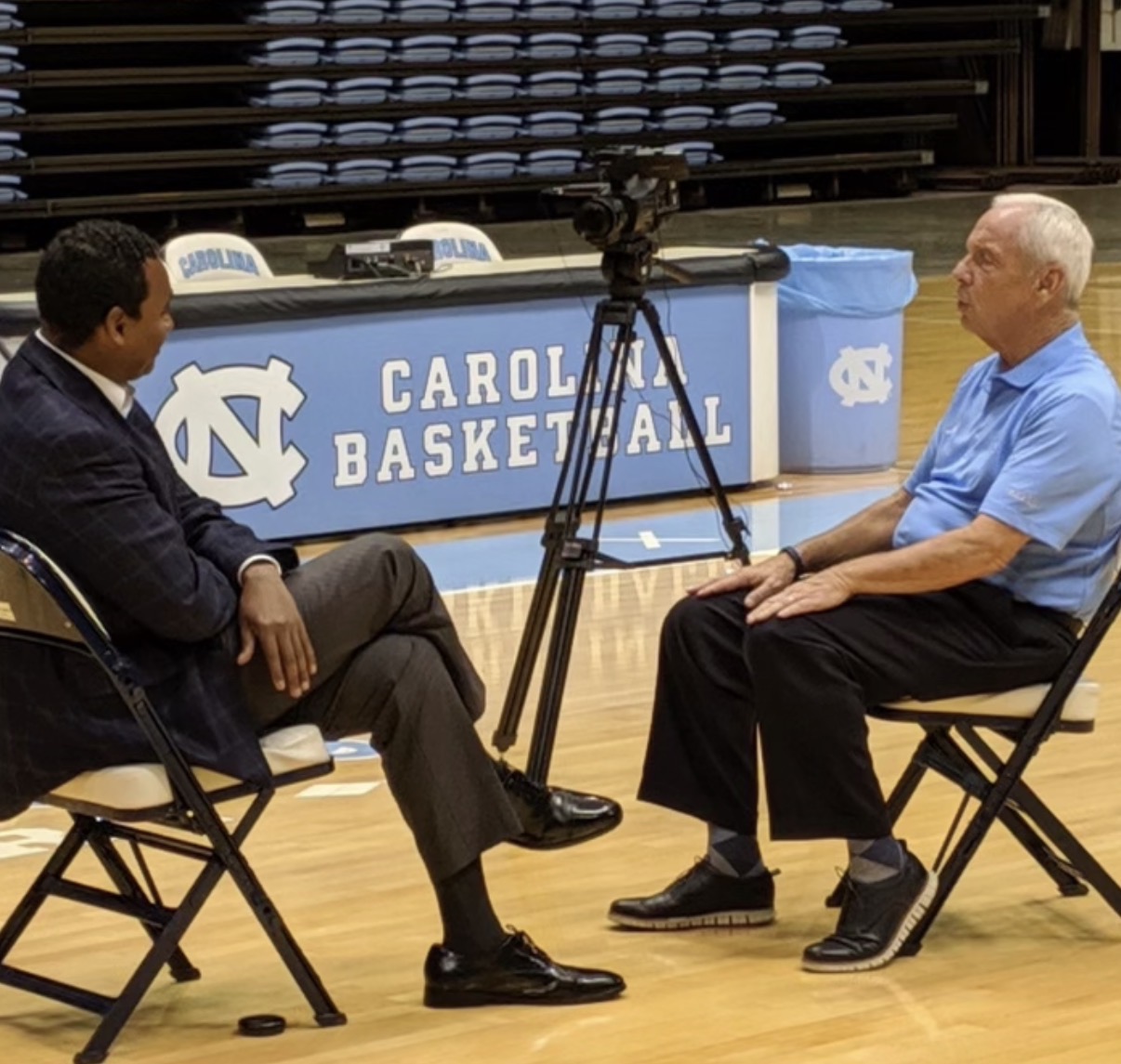 Dwayne Ballen conducting an on-camera interview on the floor at Carolina Basketball, with a cinema camera on a tripod between the two chairs