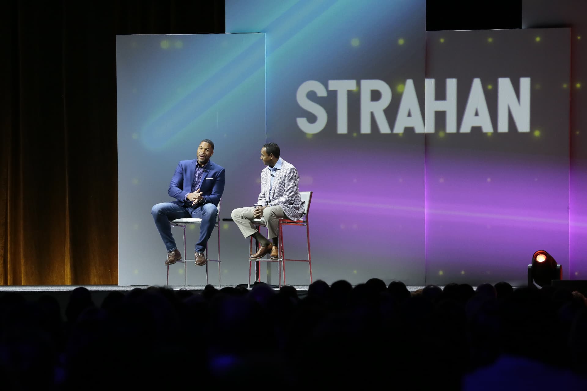 Dwayne Ballen seated on a conference main stage interviewing Michael Strahan before a large arena audience