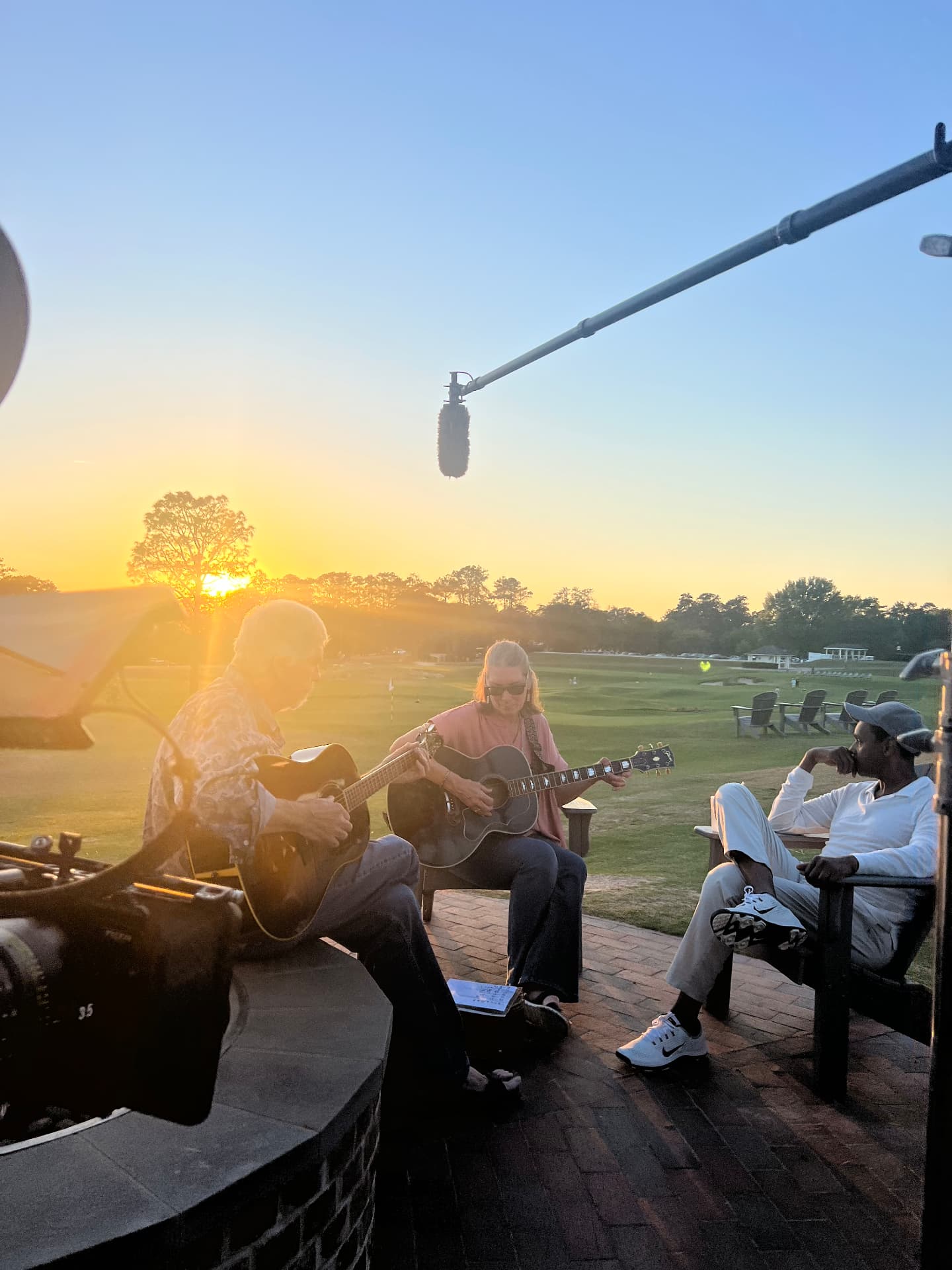 Two guitarists playing at golden hour on a golf course terrace while a boom microphone hangs over the session and Dwayne Ballen listens