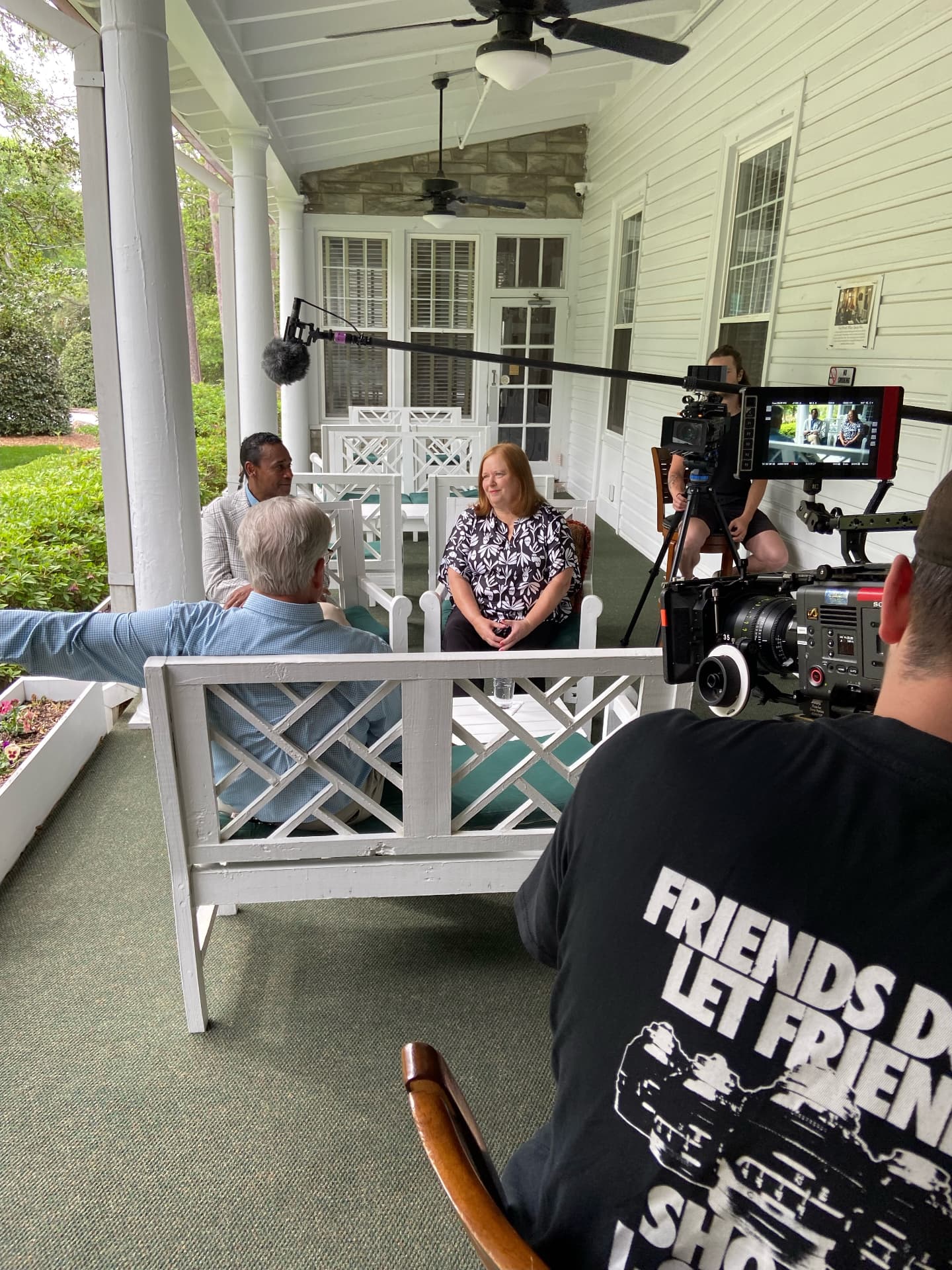A film crew rolling a cinema camera on a porch interview, with the director's monitor showing the live shot